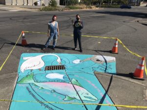 Saiyare Refaei and Gloria Joy Kazuko Muhammad celebrate the completion of the stormdrain mural they created in the McKinley neighborhood as part of the stormwater awareness campaign: “If it hits the ground, it hits the Sound”