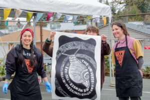 Artists hold up a large print of a snail that was made at Write253's Wayzgoose Festival using a carved and inked linoleum block and a steamroller.