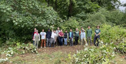 Group of volunteers stands together in a clearing surrounded by weeds that they removed during a volunteer work party