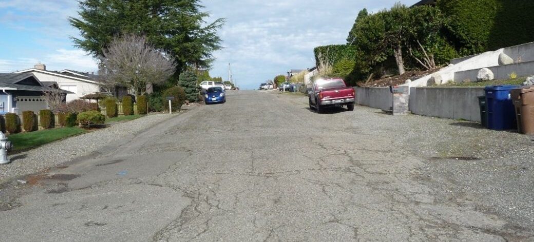 Pre-construction photo of Fir Street, taken looking north near the intersection of North 13th Street. Significant cracking and asphalt patches are visible in the pavement. Shoulders are unpaved on both sides and no sidewalks are present.