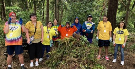 Nine volunteers stand behind a large pile of weeds, celebrating their hard work.
