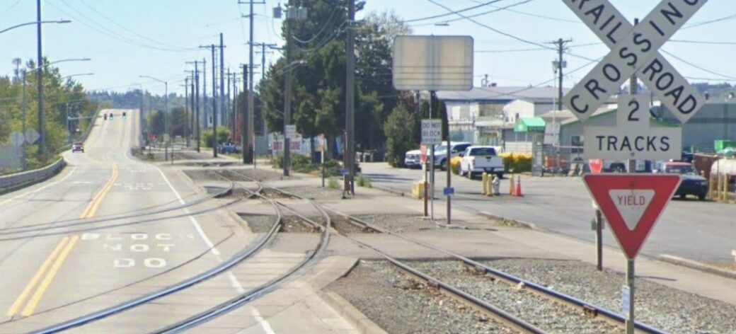 Street view of where two sets of train tracks cross Lincoln Avenue, approximately 900 feet away from Port of Tacoma Road. It shows the pre-construction rail crossing infrastructure here: a sign post with a 