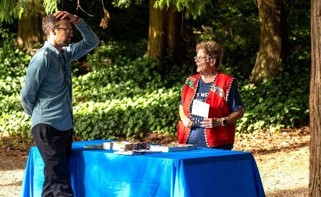 Photo of City staff tabling at an outdoor community event, with a staff member in a red vest speaking with a community member across a table covered in a blue cloth, surrounded by trees.